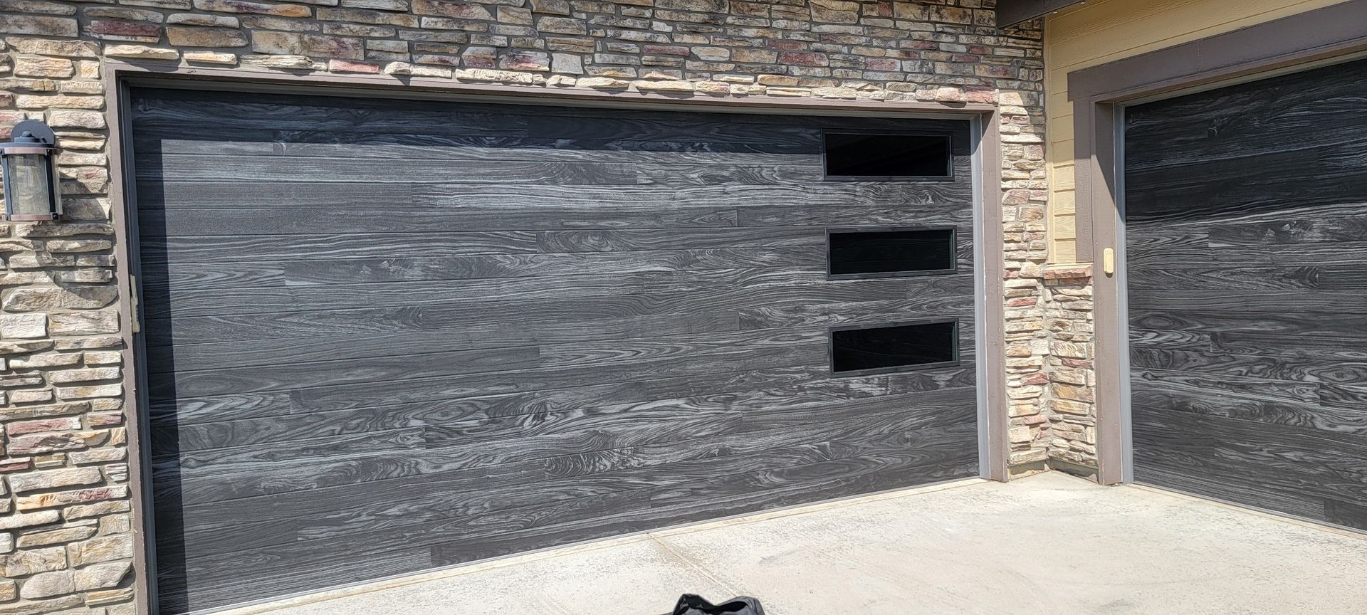 Garage door with dark gray wood-like panels and rectangular windows, set in a stone facade.