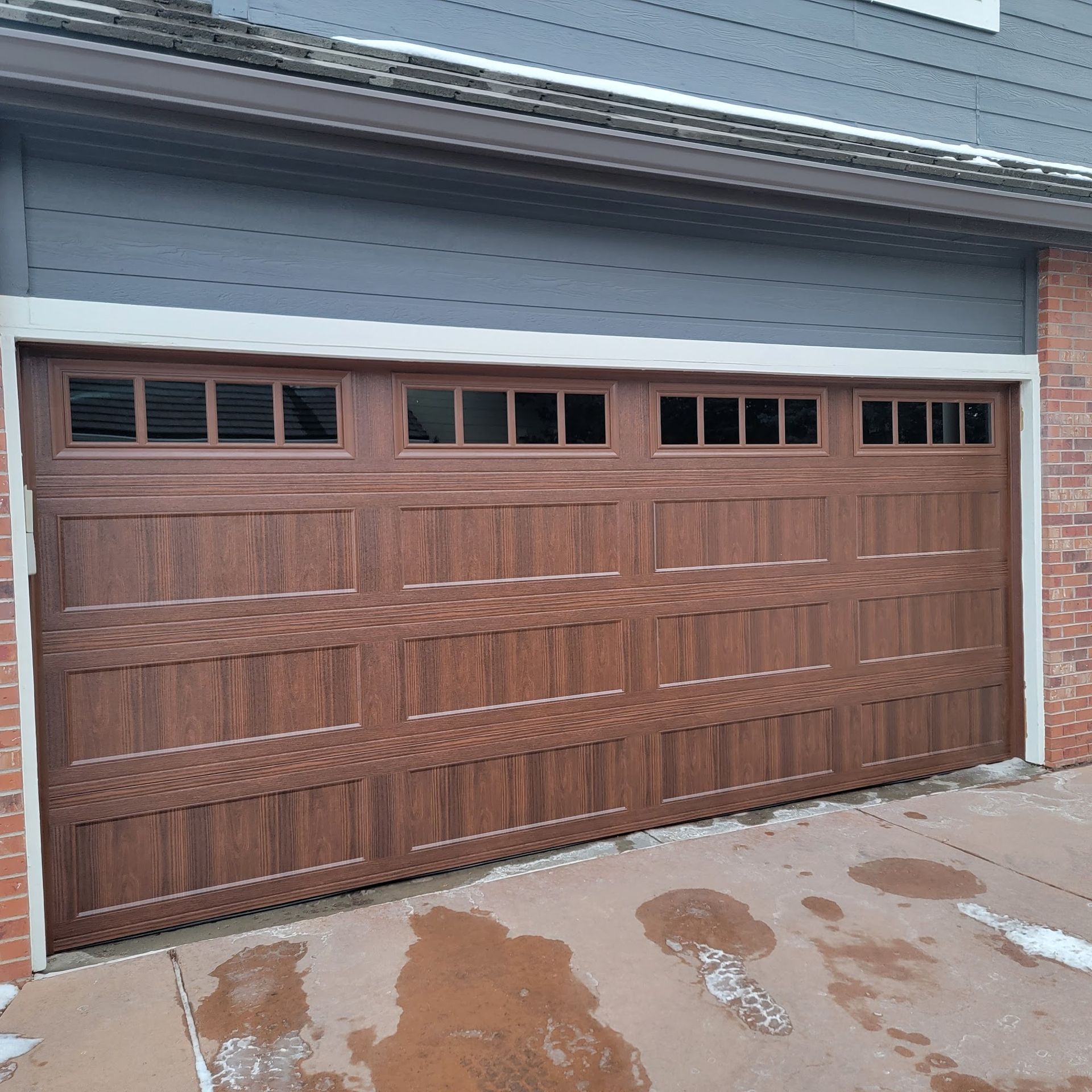 Brown wood-grain garage door with windows at top, set into brick and blue siding.