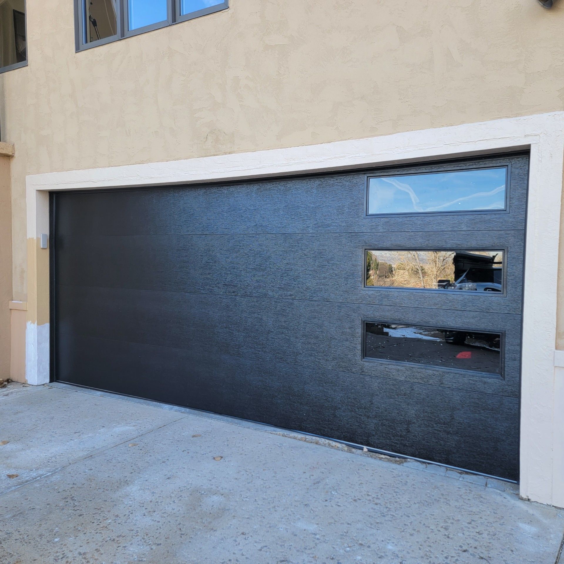 Dark gray garage door with three rectangular windows; the house is beige.