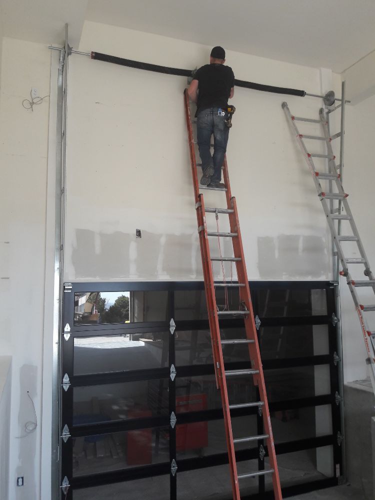 Man on ladder installing garage door spring above a partially installed glass door.