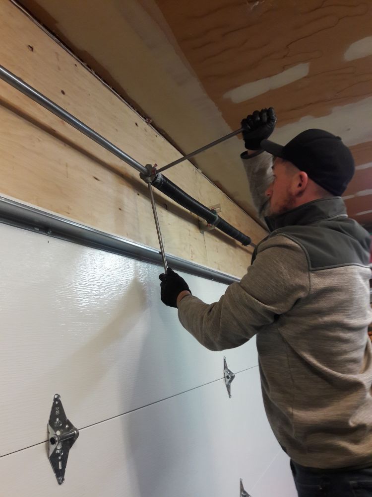Man in gloves and hat tightening a garage door spring with a winding bar.