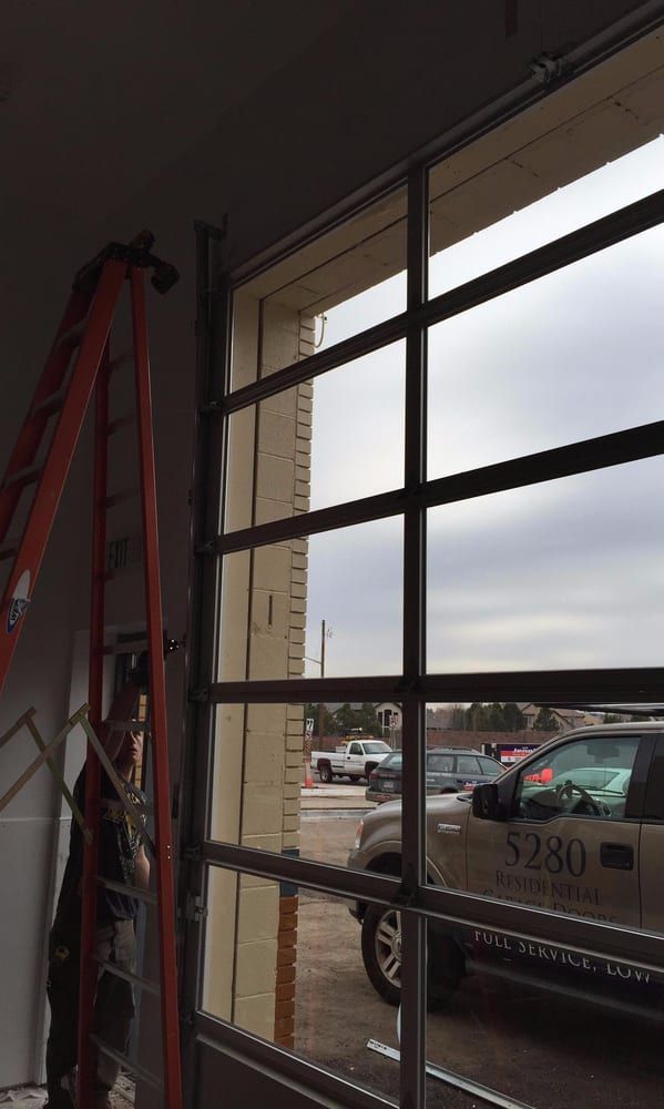 A partially installed glass garage door with a ladder inside. A cloudy day is visible through the glass panes.