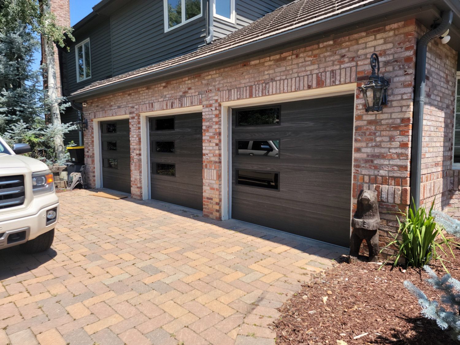 Three dark garage doors with glass panels on a brick building. A truck is parked in the brick driveway.