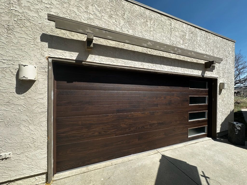Dark brown garage door with three rectangular windows, on a stucco building, under a pergola.