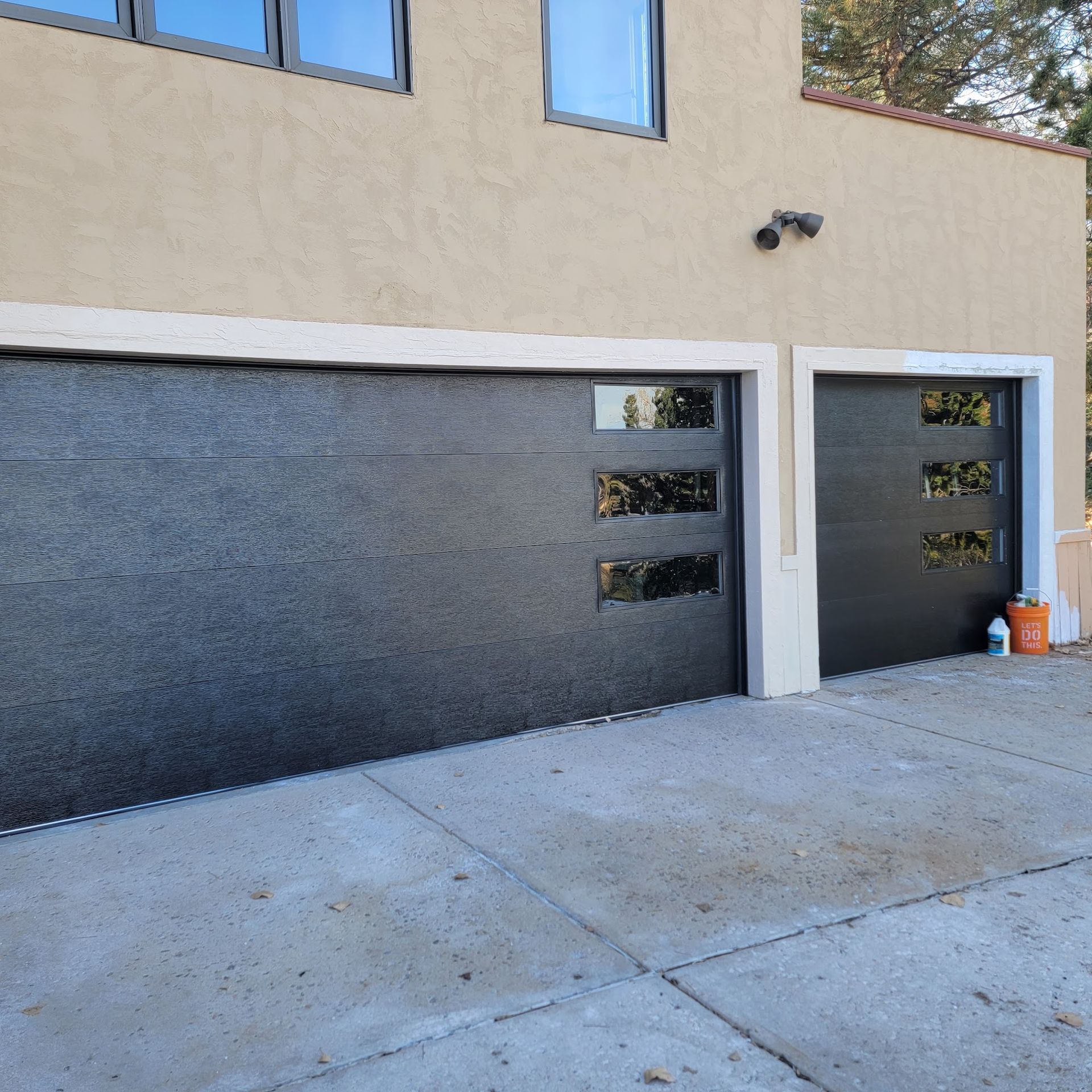 Two black modern garage doors with glass windows on a concrete driveway, beige building.