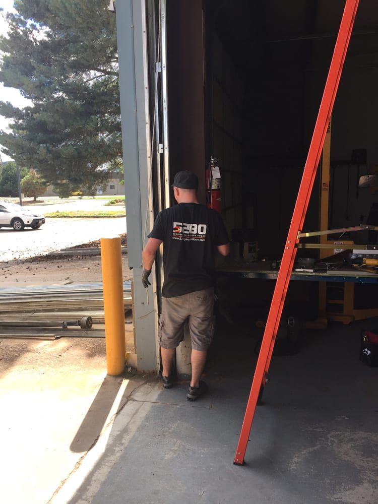 Man in black shirt and shorts walking into a dark shop with a partially open door and orange ladder.