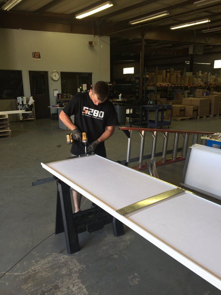 A man uses a drill on a large white board with a metal strip in a workshop.