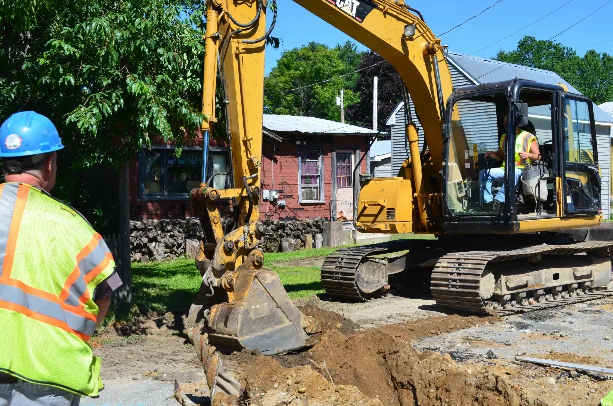 A man in a hard hat stands in front of an excavator.