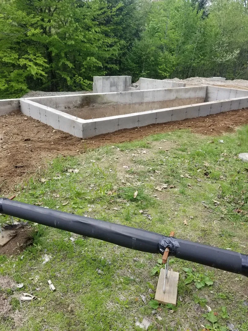 A black pipe is sitting in the grass next to a concrete foundation.