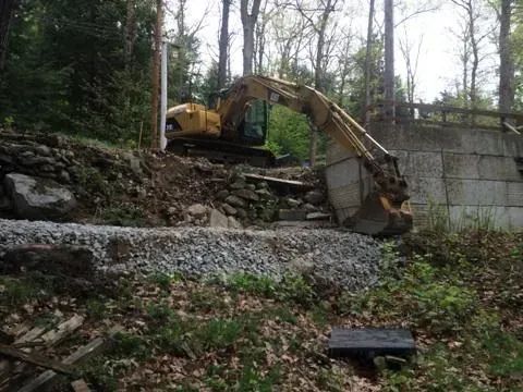 An excavator is working on a rock wall in the woods.