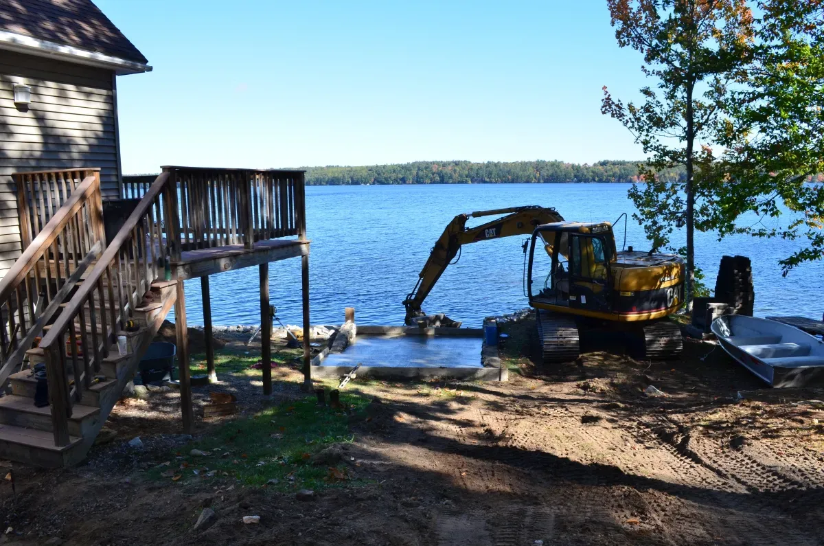 A large yellow excavator is sitting on the shore of a lake next to a house.