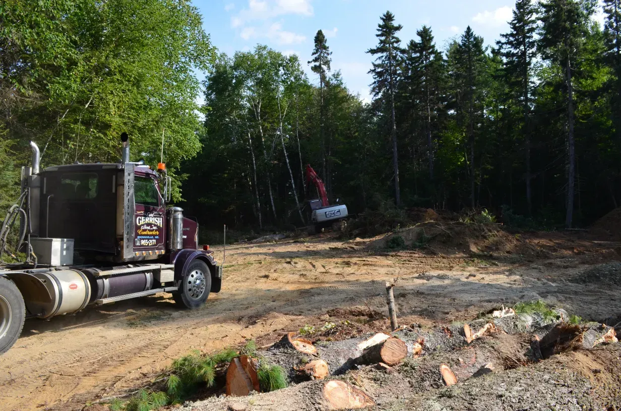 A semi truck is driving down a dirt road in the middle of a forest.
