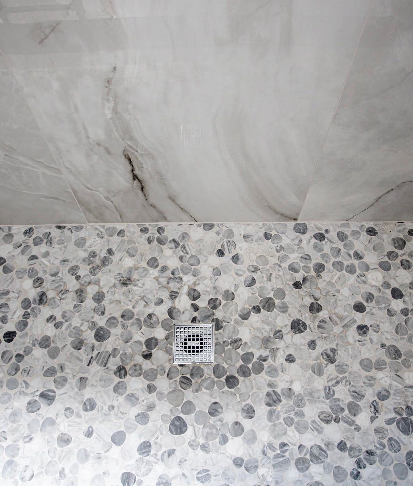 Bathroom wall with gray marble tile above pebble-patterned tile and a small square drain grate