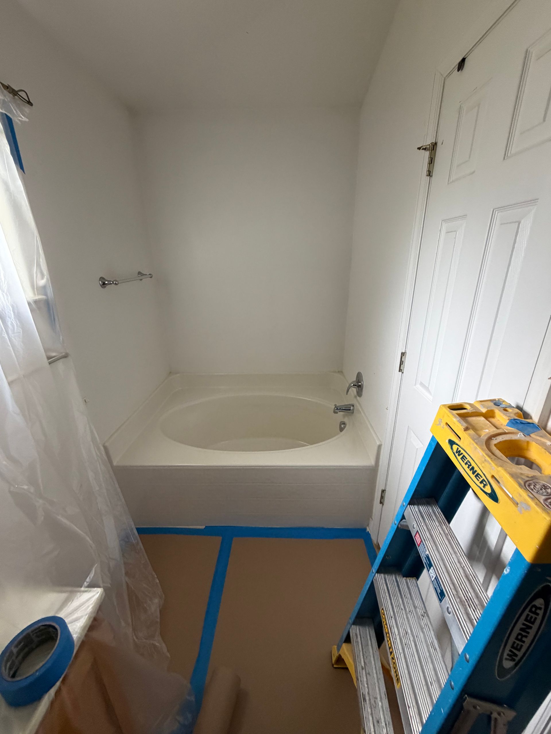 Small bathroom under renovation with tub, blue painter’s tape, and a ladder-like shelf of supplies.