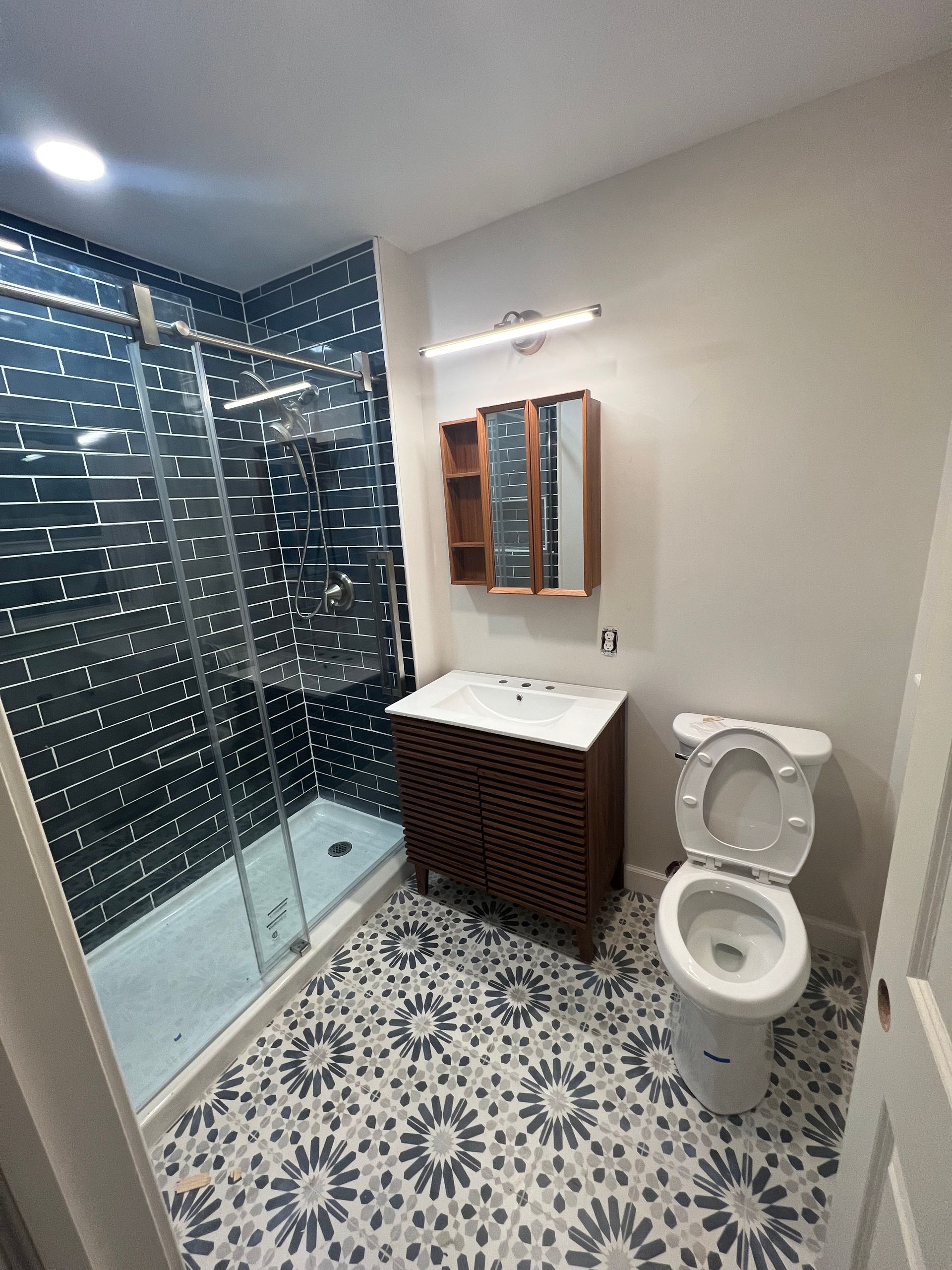 Modern bathroom with glass shower, patterned tile floor, sink cabinet, mirror, and toilet.