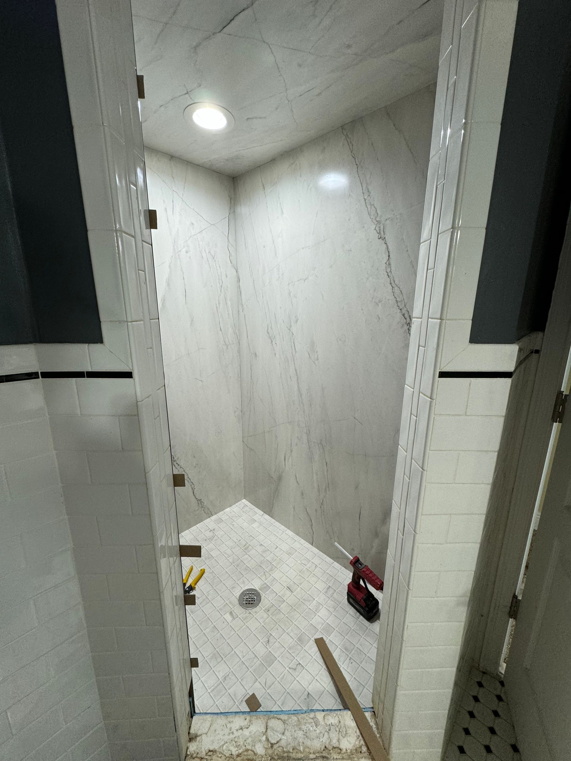 Narrow tiled shower stall with white walls, ceiling lights, and cleaning items on the floor.