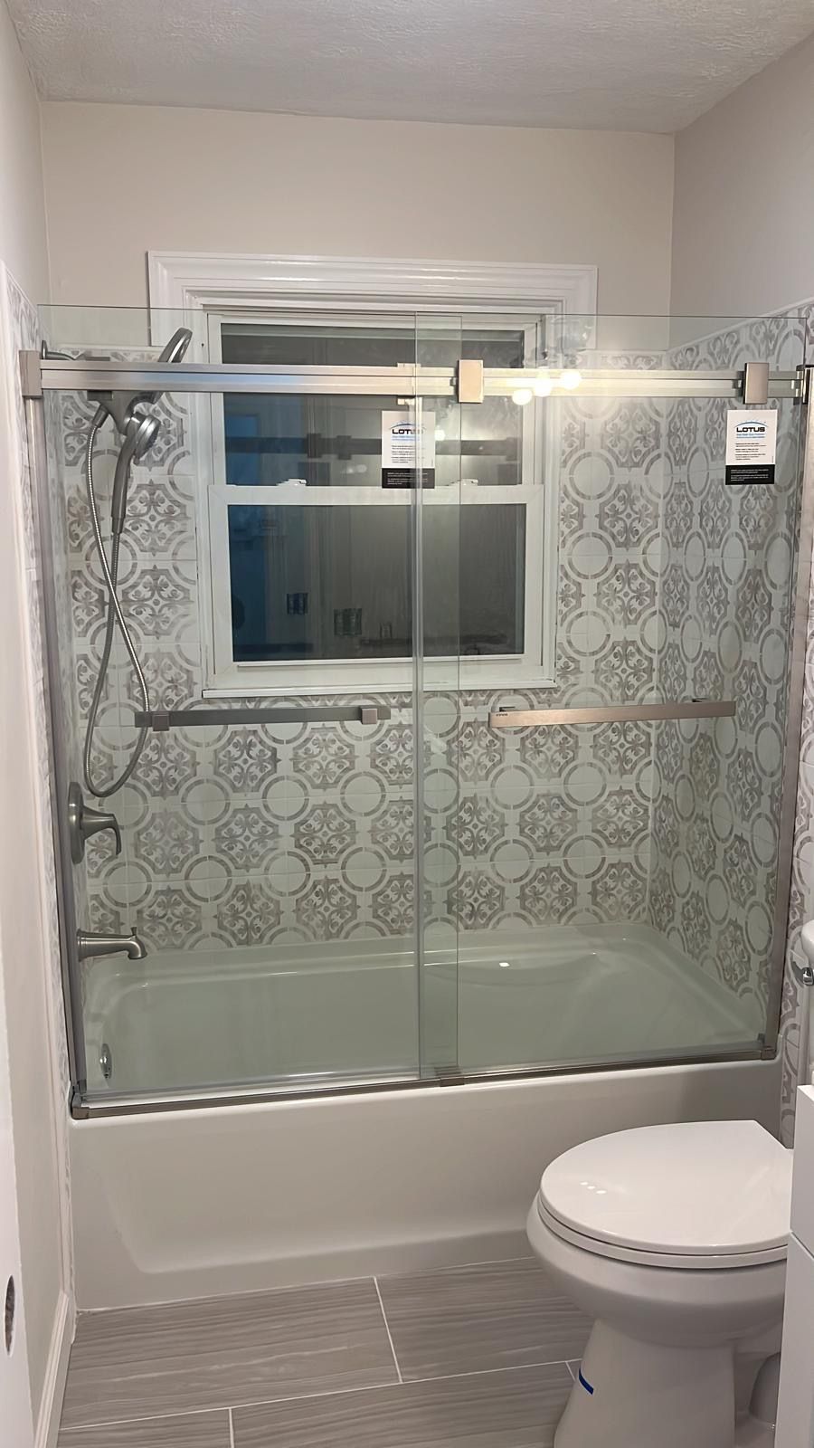 Bathroom with a glass shower-tub combo, patterned tile walls, window, and a toilet in the foreground