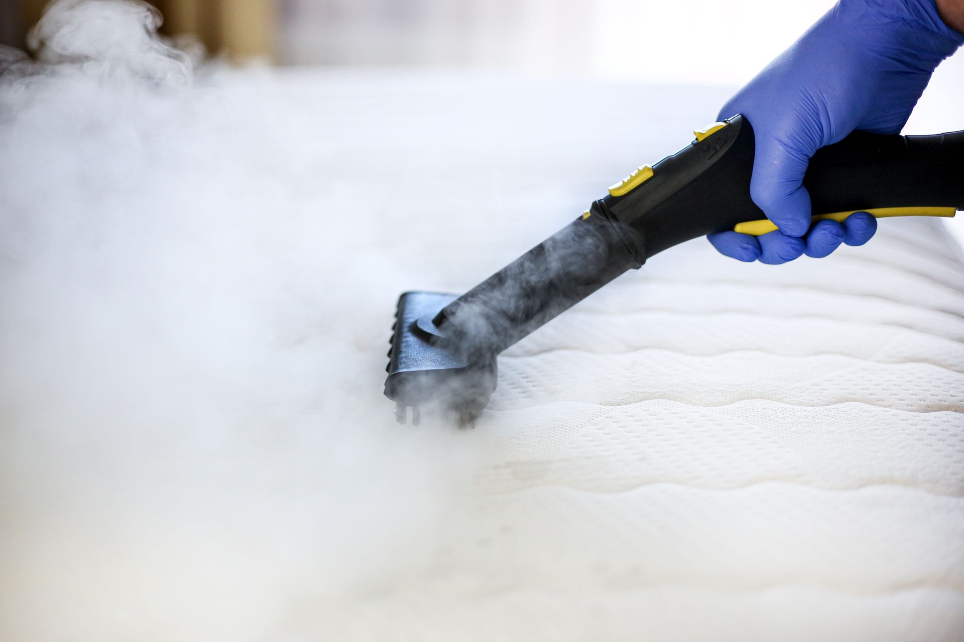 Person wearing blue gloves using a steam cleaner on a white mattress. Steam is visible.