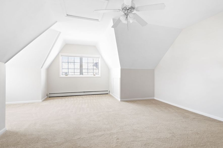 Empty attic room with sloped white walls, carpet, and window.