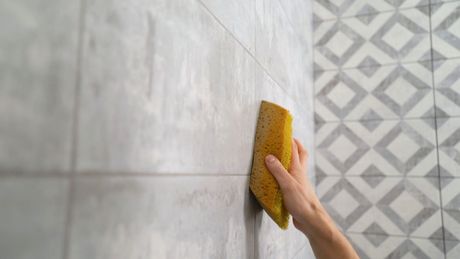Person cleaning light gray bathroom tiles with a yellow sponge.