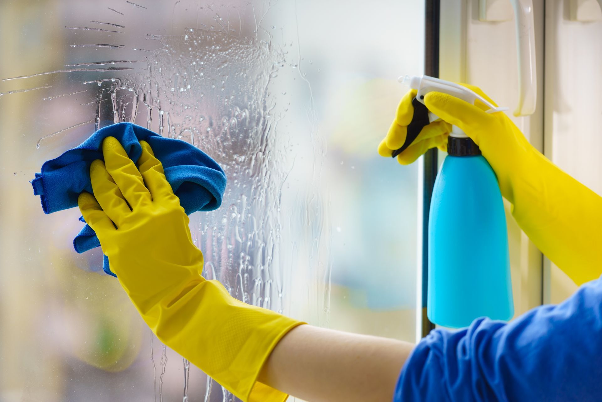 Person in yellow gloves cleaning window with spray bottle and blue cloth.