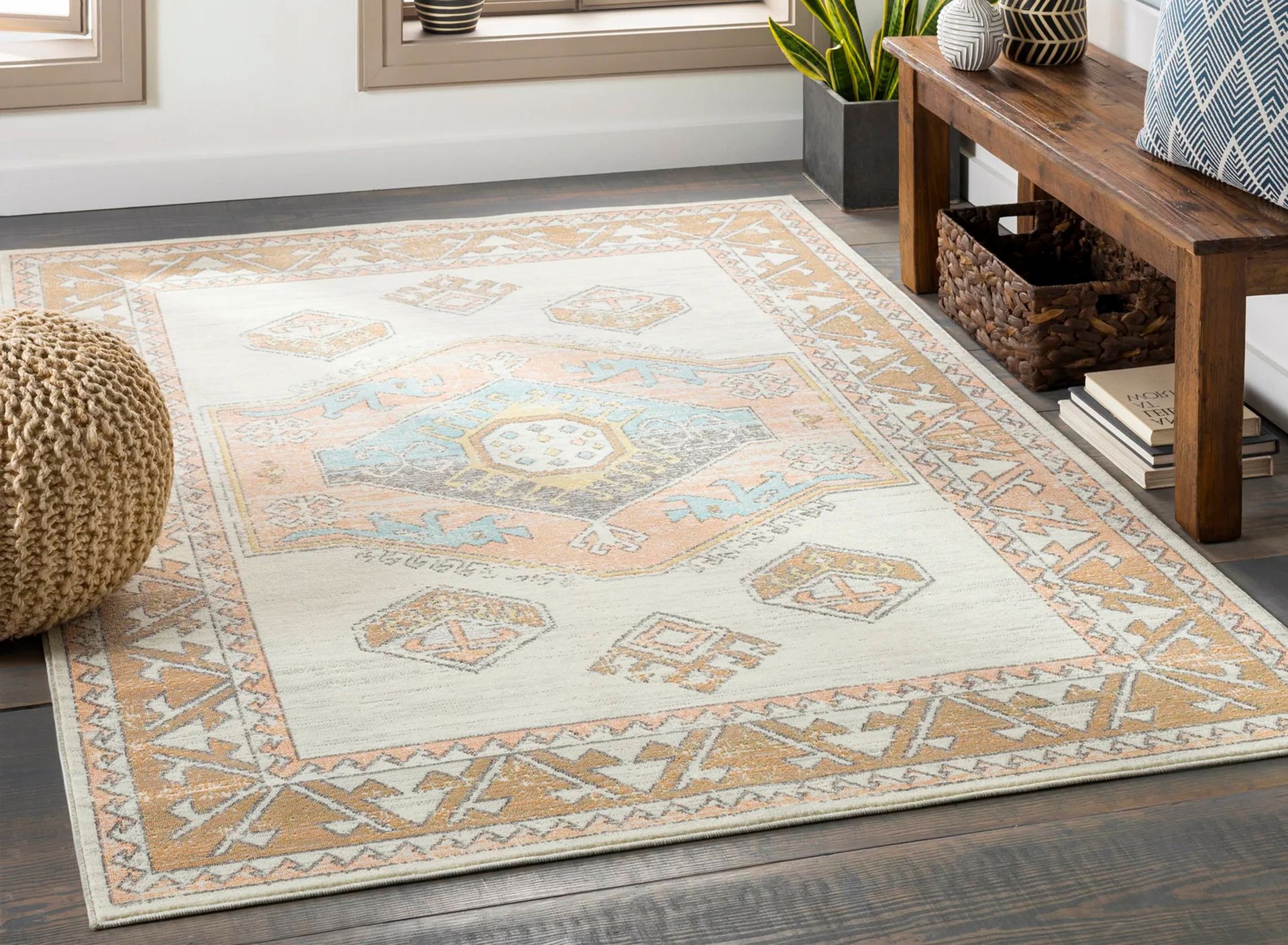 Beige and peach patterned rug on wooden floor. A bench, ottoman, and plants are nearby.
