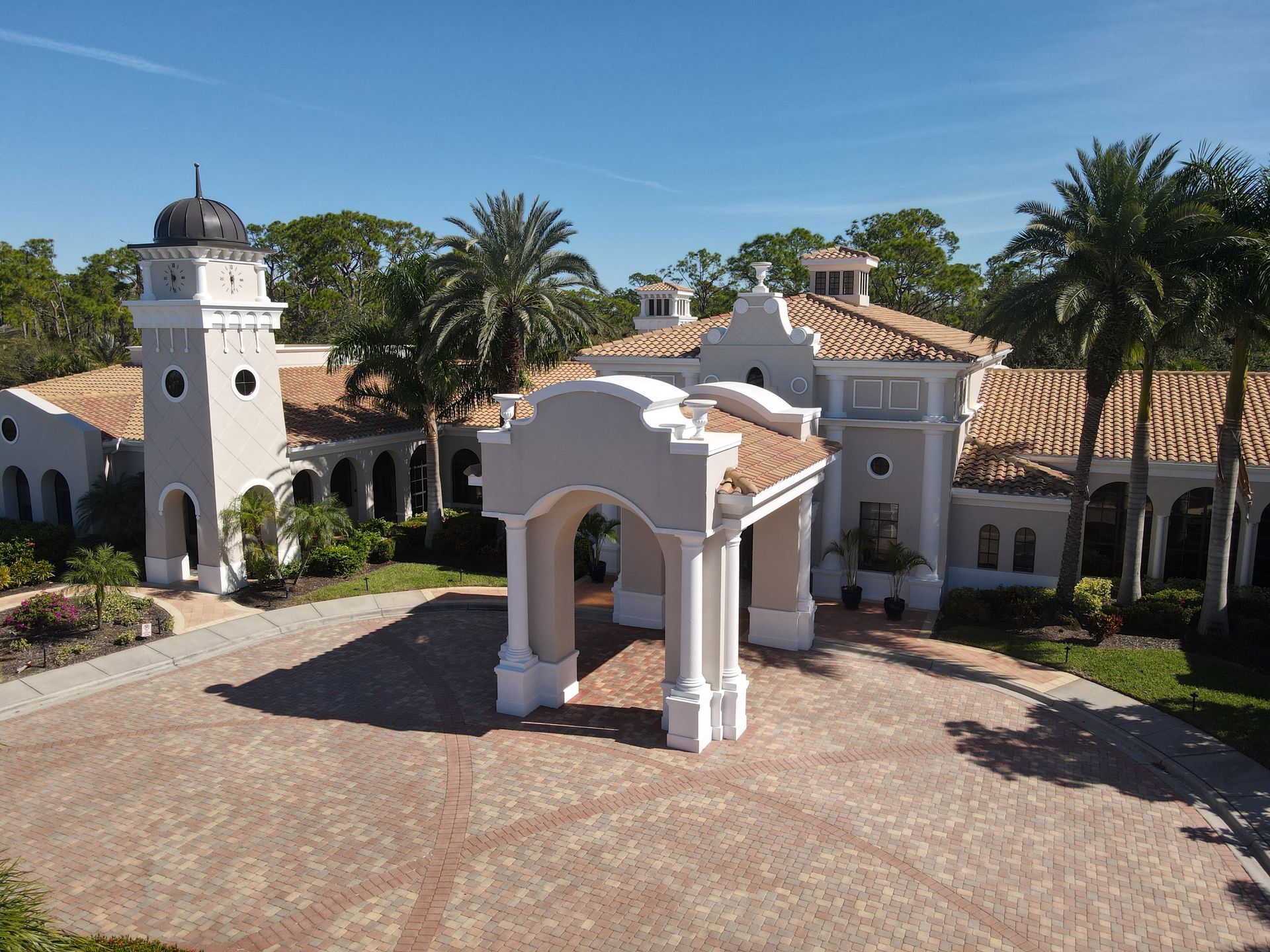 Mansion with light-colored walls, red tile roof, and circular driveway, under a bright blue sky.