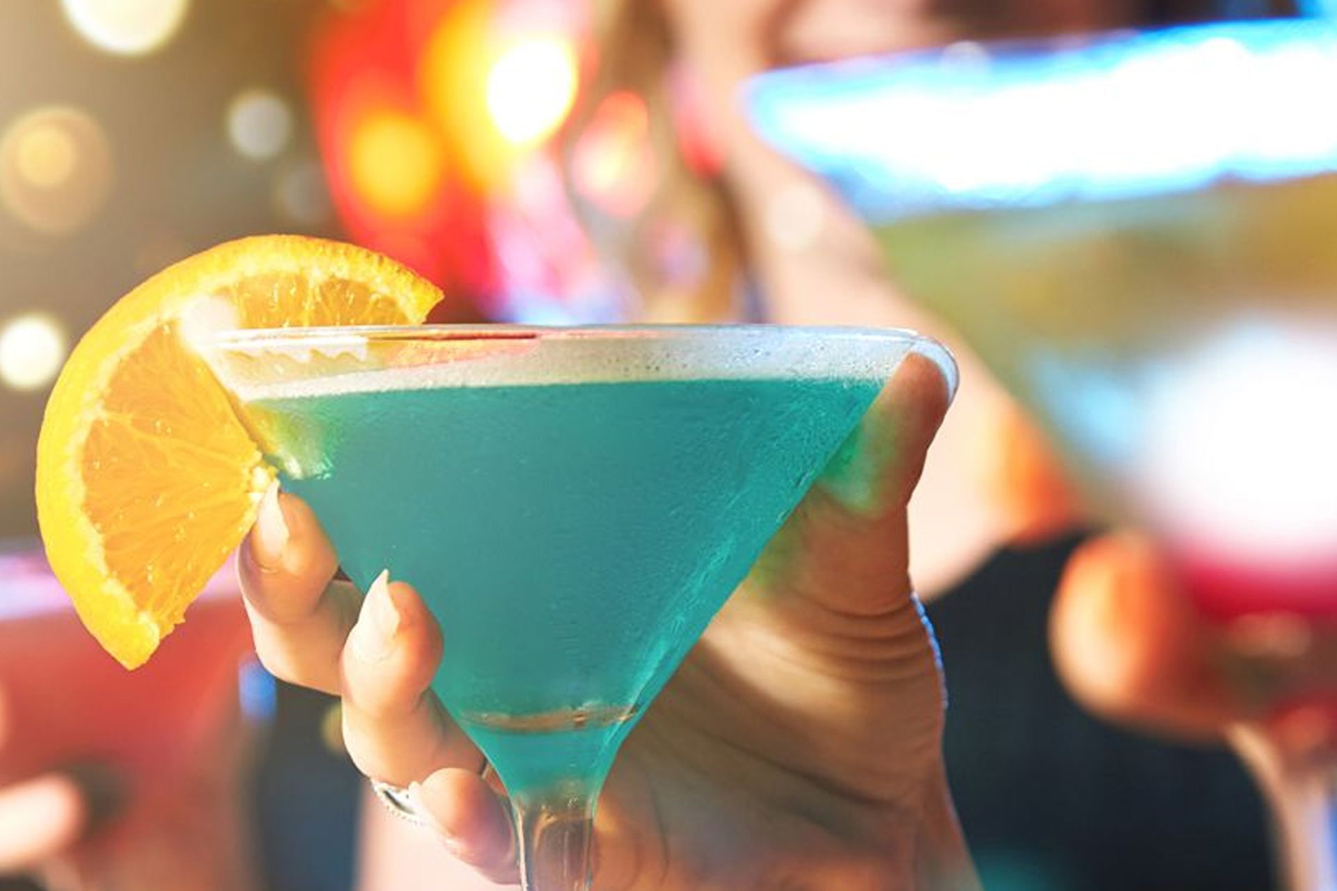 Hand holding a blue cocktail with orange slice, blurred background with other drinks.
