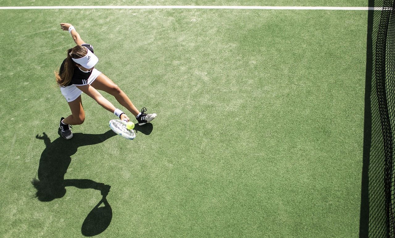 Tennis player in white shorts and visor swings racket on green court, shadow visible.