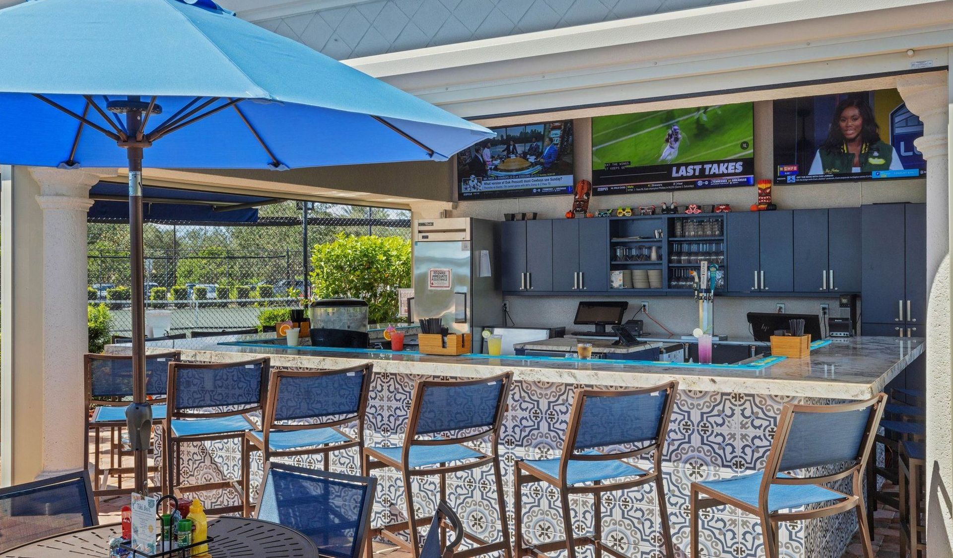 Outdoor bar with blue umbrella and stools, TV screens, and a long countertop.