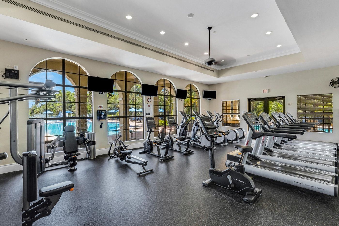Gym with exercise machines, large windows overlooking a pool, and black flooring.
