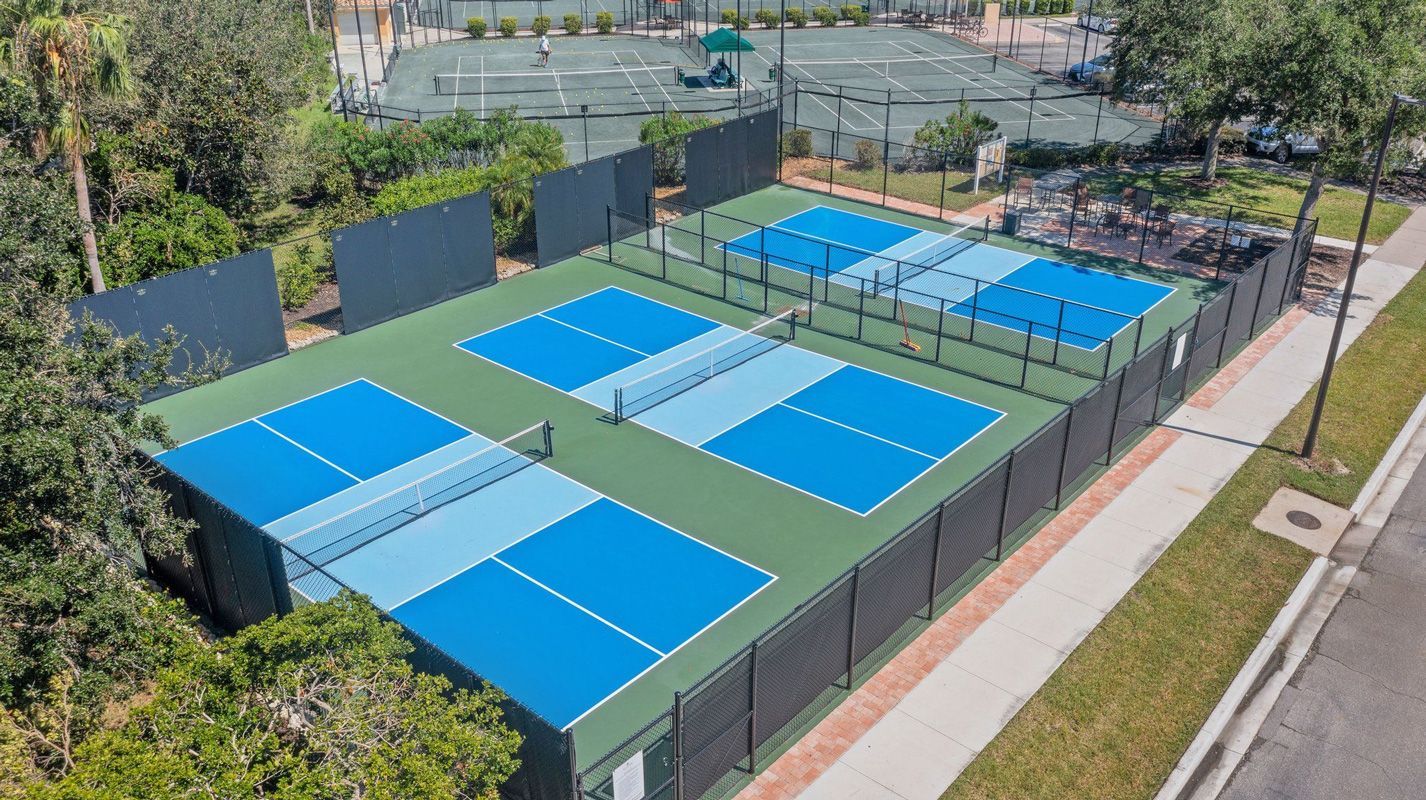 Overhead view of several blue pickleball courts enclosed by black fencing, in an outdoor setting.