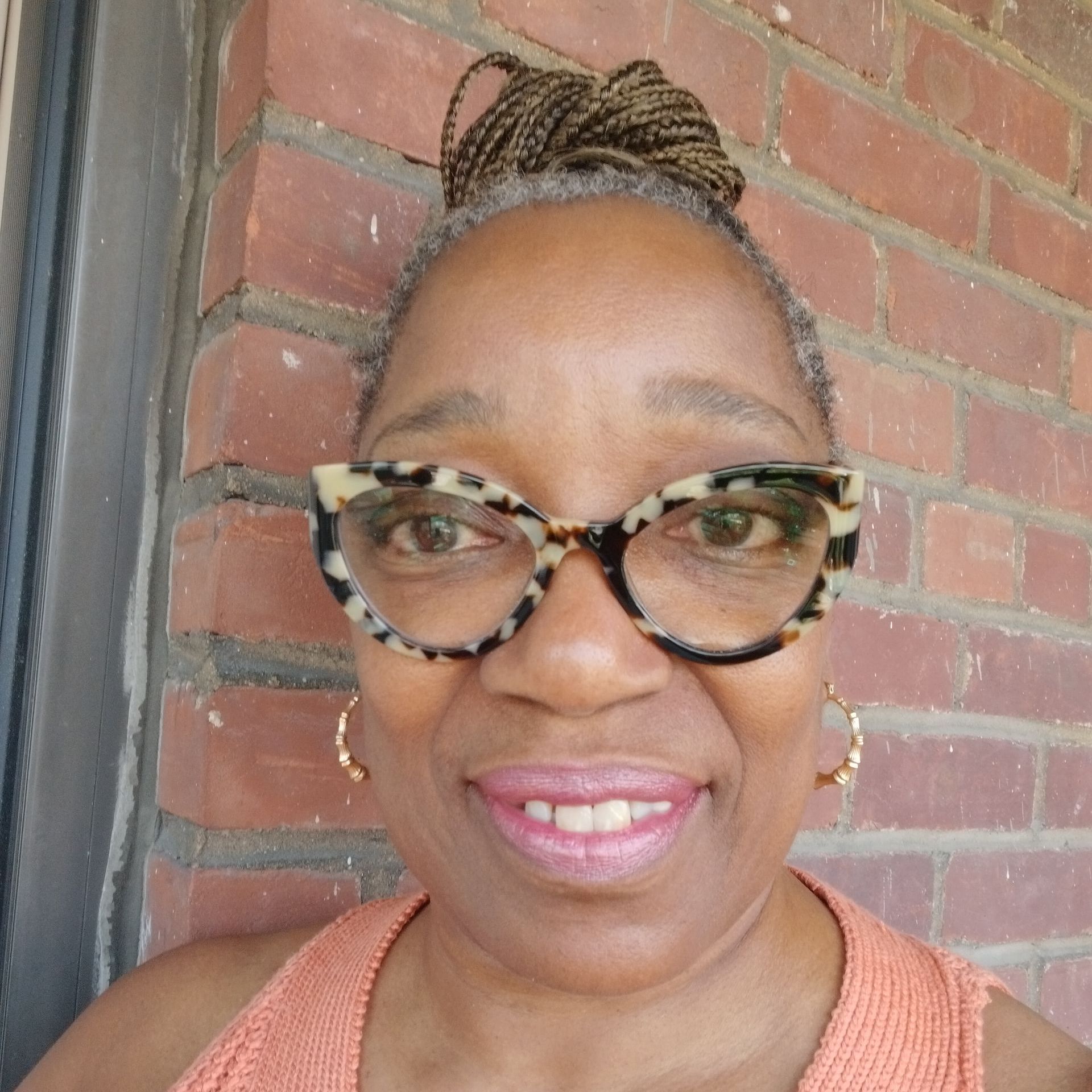 A woman wearing glasses and earrings is smiling in front of a brick wall.