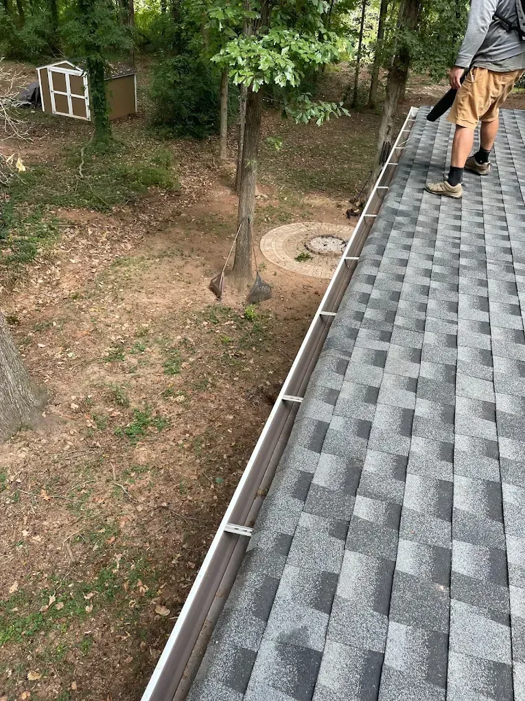 Person cleaning roof gutters with a blower. Brown shingles, white gutters, and trees in a yard.
