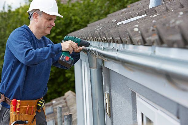 Man in hard hat installing gutter with power drill on a house roof.