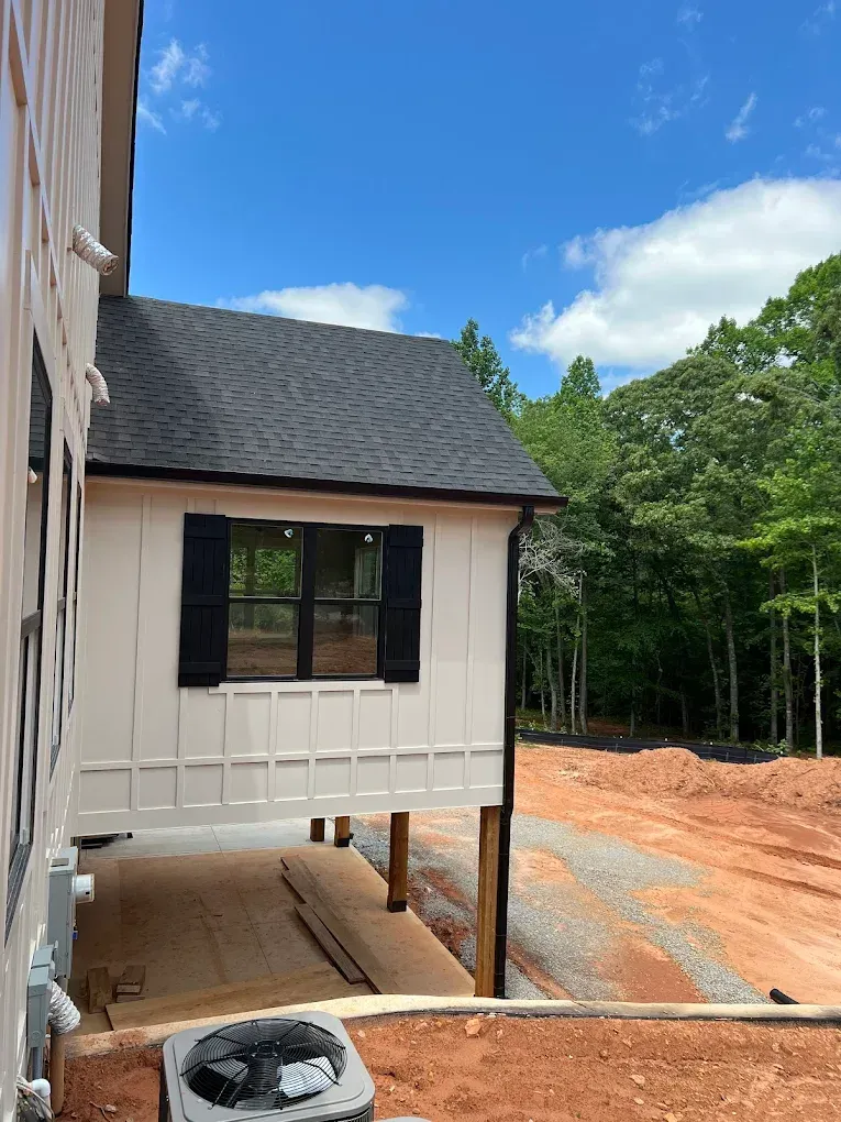 Exterior of a house under construction; white siding, black shutters, black roof, and dirt ground.