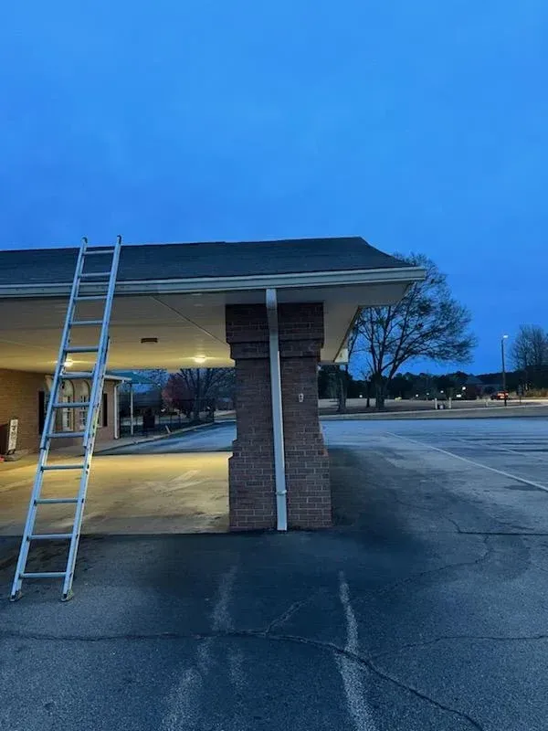 Ladder leaning against a building's roof; a white downspout runs down a brick pillar. Dusk setting with a blue sky.