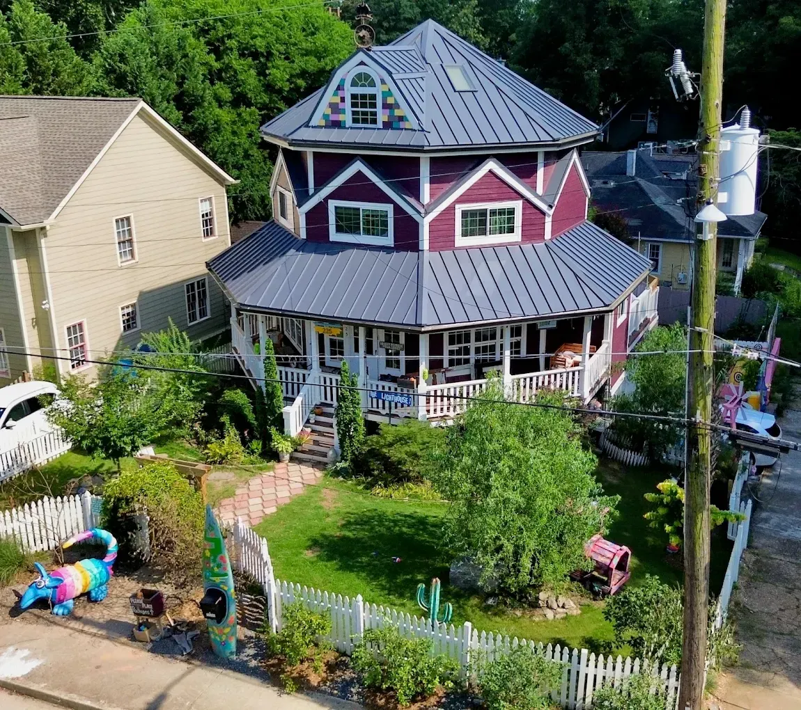 A unique, multi-story round house with a wraparound porch, purple siding, and gray roof, surrounded by a white picket fence.