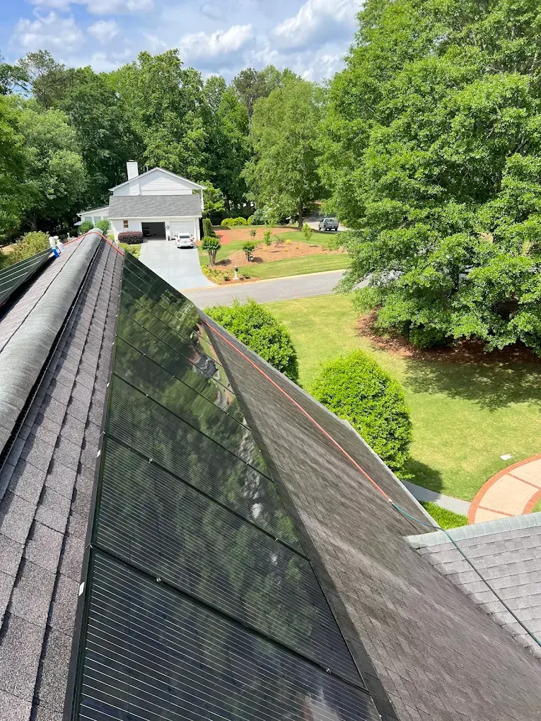 Solar panels on a dark gray roof. In the background is a house, driveway, and trees under a blue sky.