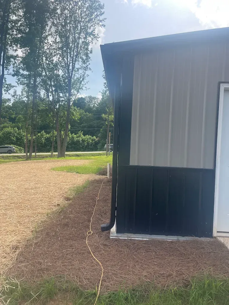 Black and gray metal building with a gutter and brown mulch. Green grass and trees in the background.