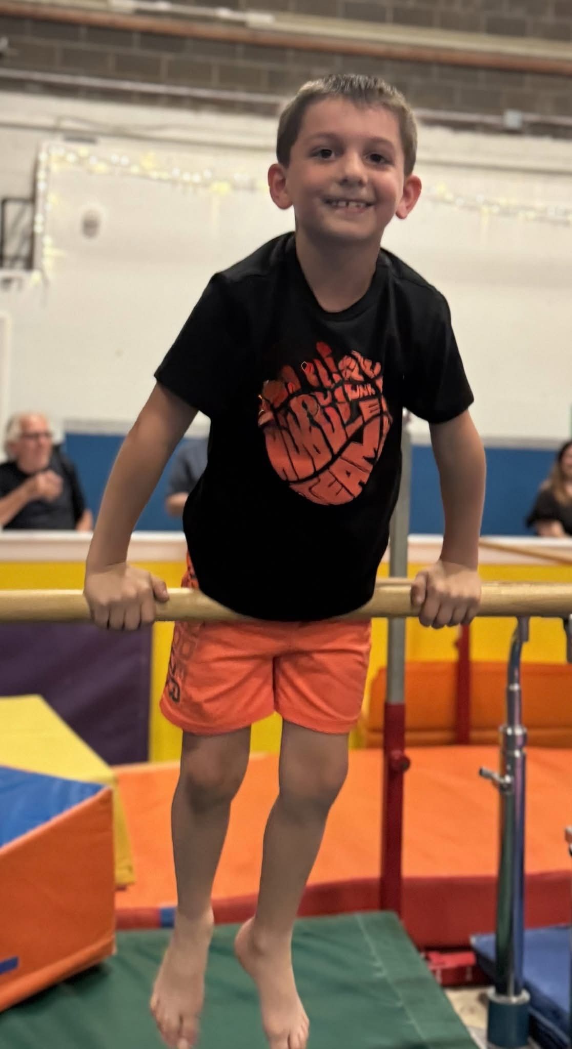 Boy smiling, hanging from parallel bars in a gym. He wears orange shorts and a black shirt.