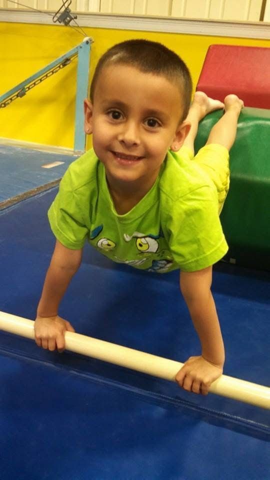 Boy in green shirt smiling, gripping a horizontal bar, legs raised, in a gym.