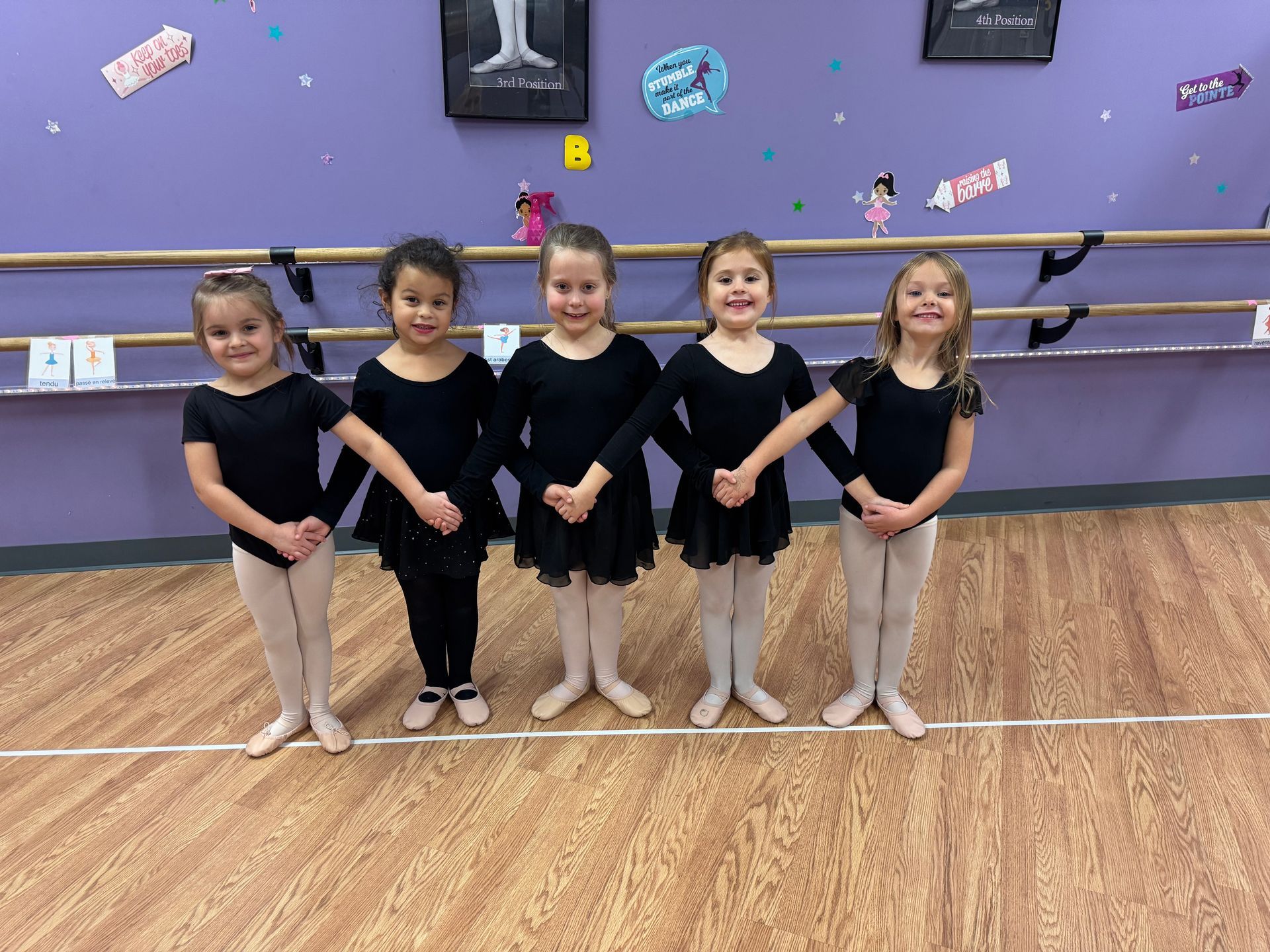 Five young girls in black leotards and ballet shoes stand in a dance studio, holding hands, smiling.