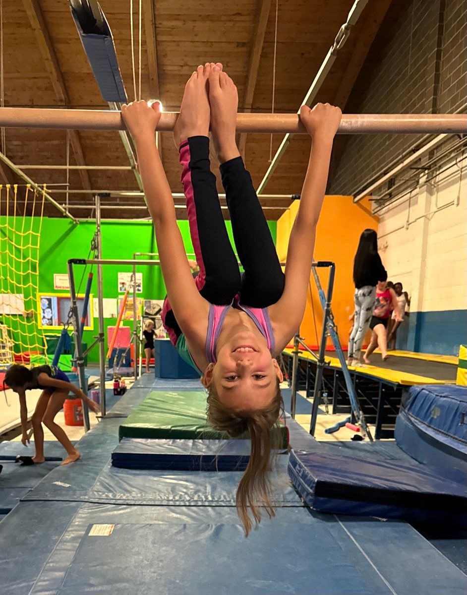 Girl hanging upside down on a bar in a gymnastics gym, smiling.