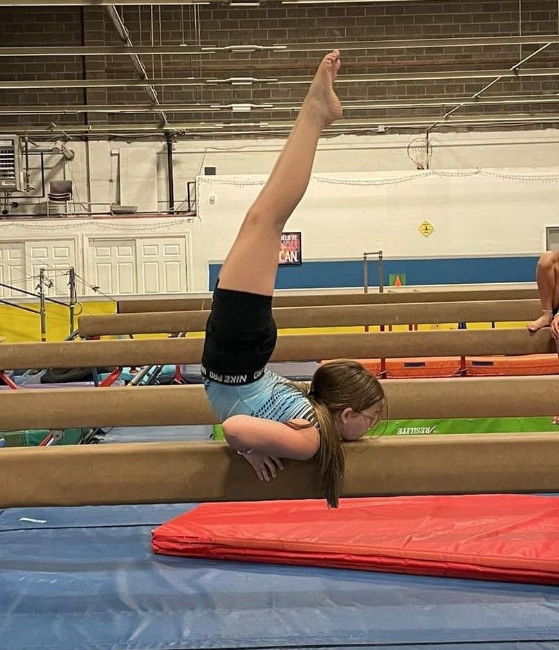 Gymnast balances upside down on a beam, legs extended, with hands on the beam. Indoors.