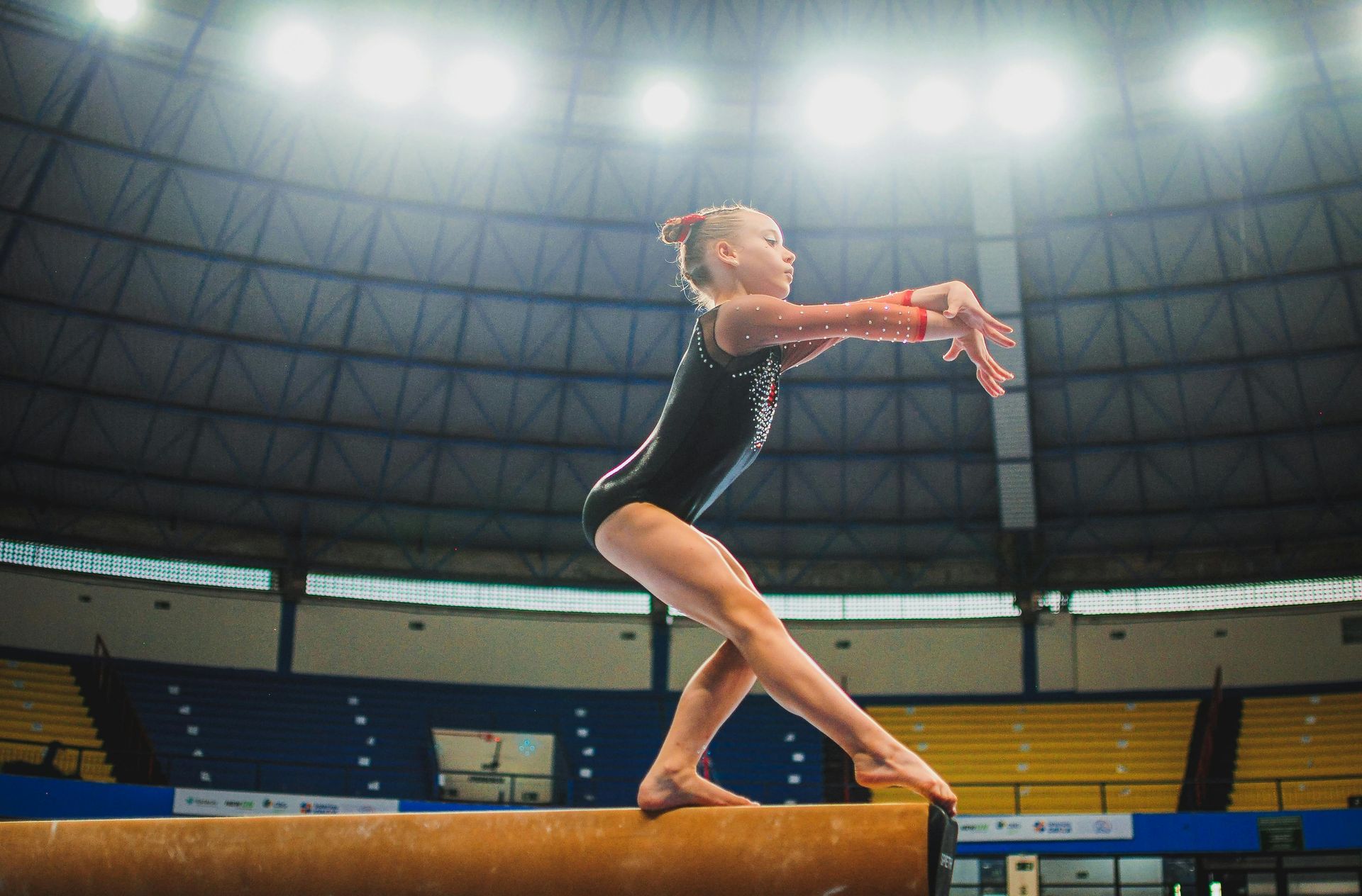 Gymnast balances on a beam, arms extended, in a gymnasium under bright lights.