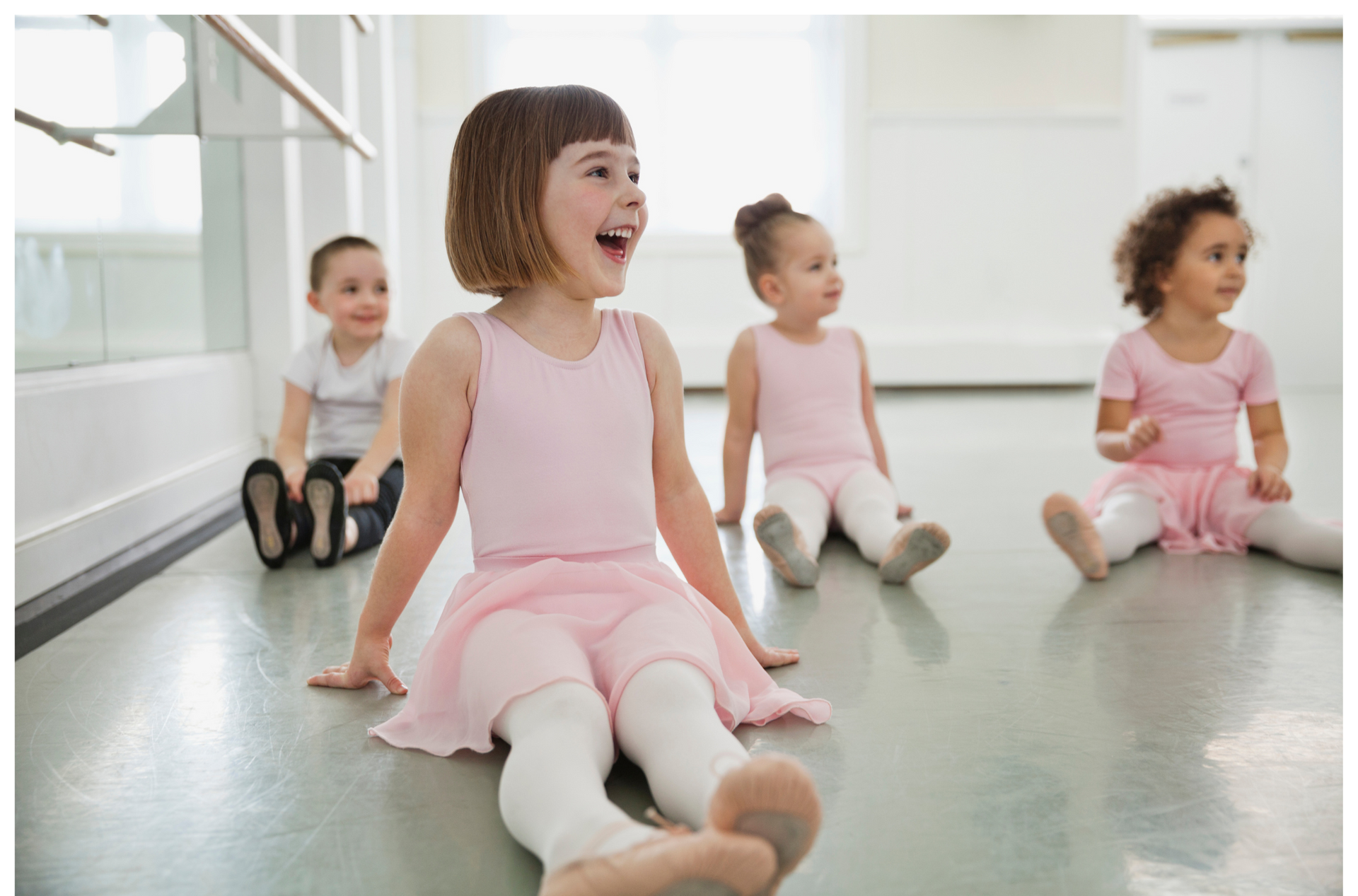 Children in pink ballet attire sitting on a dance floor, one laughing.