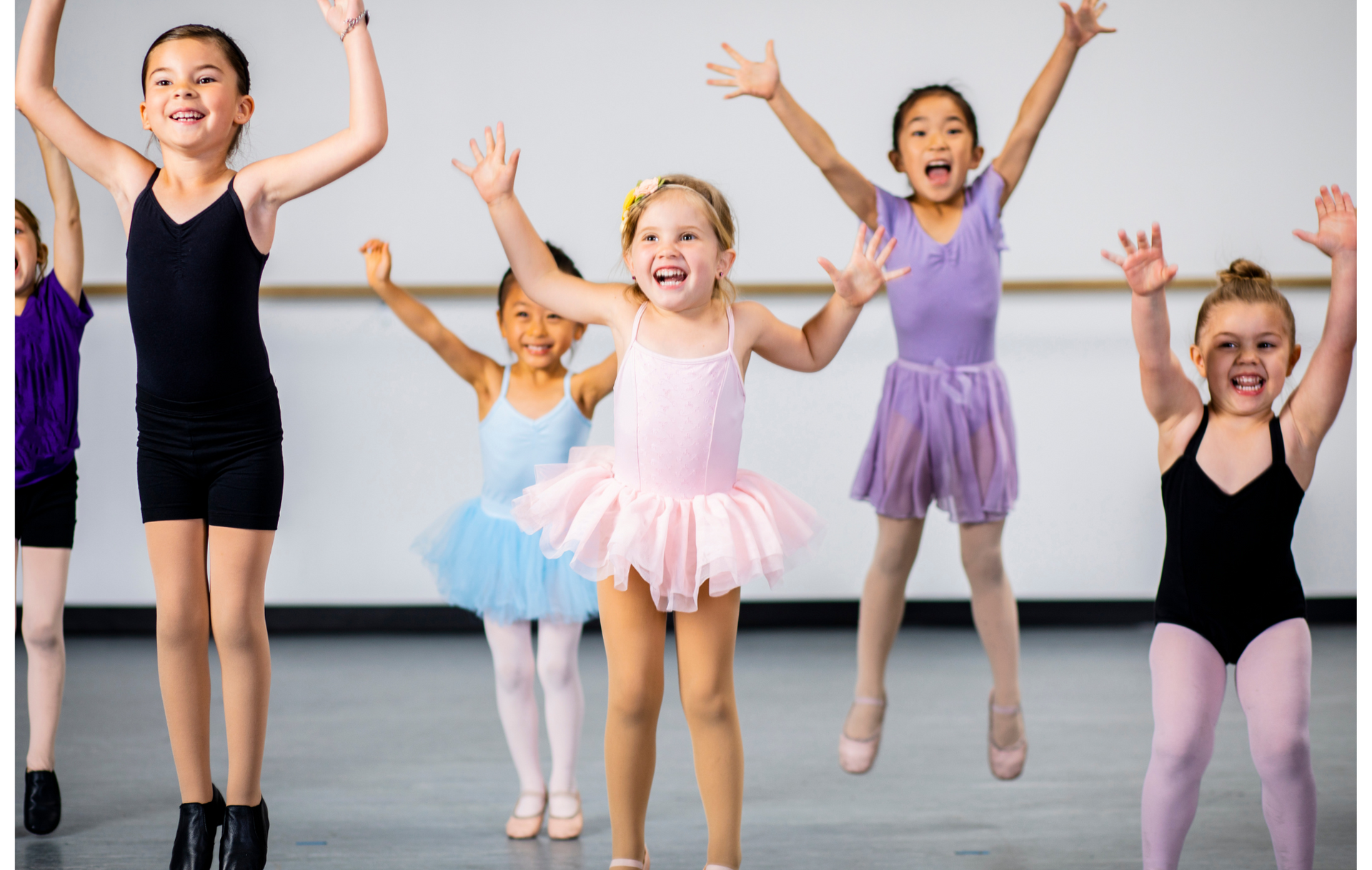 Children in dance class jumping, arms raised, smiling. Wearing leotards and tutus. Dance studio.