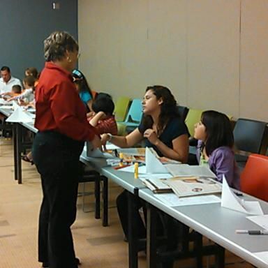 Woman in red shirt talking to seated people at a table; others work on crafts in a room.