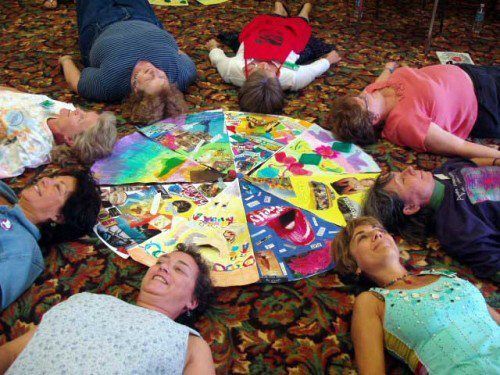 Group of people lying around a colorful artwork on a patterned carpet, looking upward.